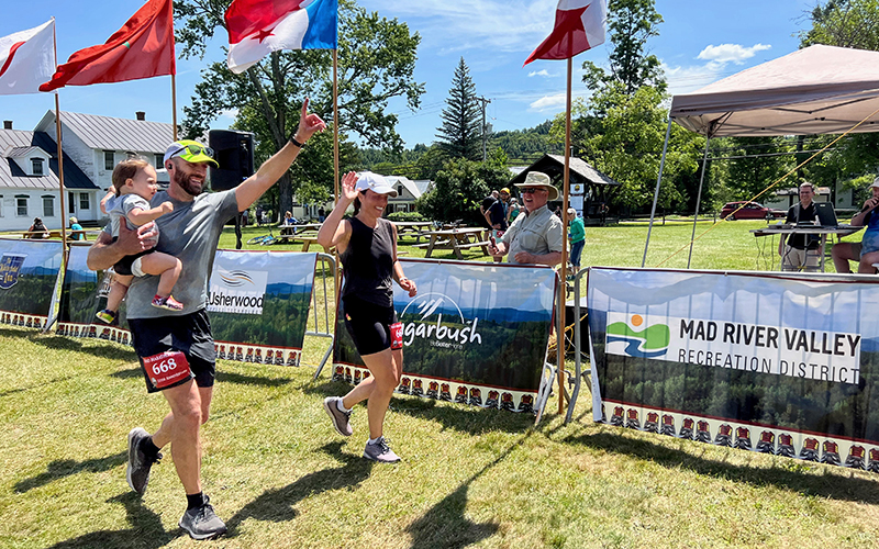A family of runners with dad carrying a baby cross the finish line of the 2022 Mad Marathon in Waitsfield, VT. Photo: Bear Simmons