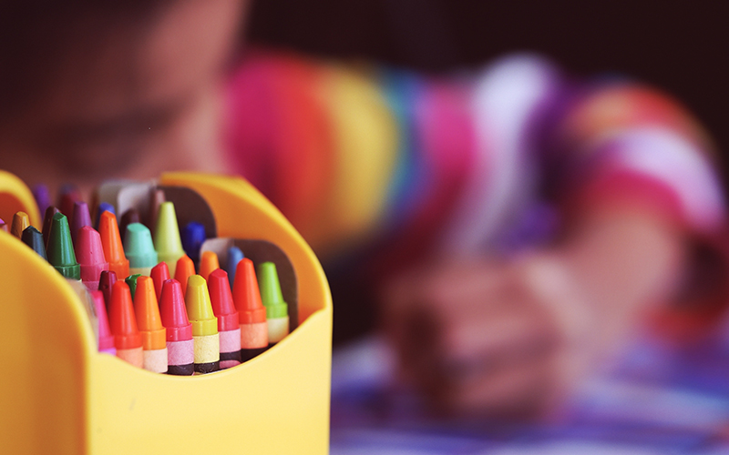 crayons in the foreground with a child coloring in the background.