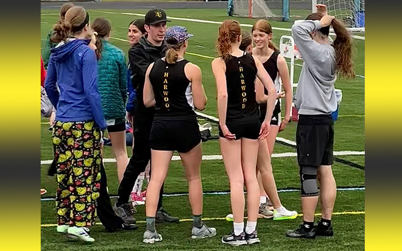 Harwood track coach Jake Pitman with relay team members (left to right) Heidi Haraldsen (purple hat), Julia Cisz (red head with pony tail), Celia Wing (in front of Cisz), and Adleigh Franke (facing camera) as they prepare for the girls 4x800 relay team.